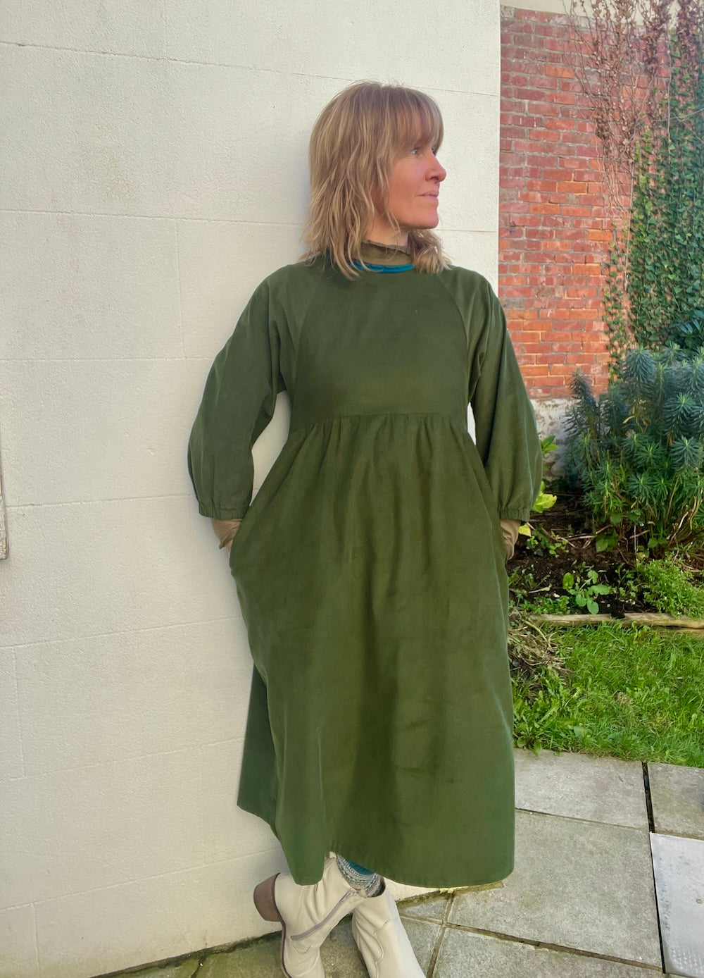 Model wearing a mossy green needlecord dress with hands in pockets looking in the distance
